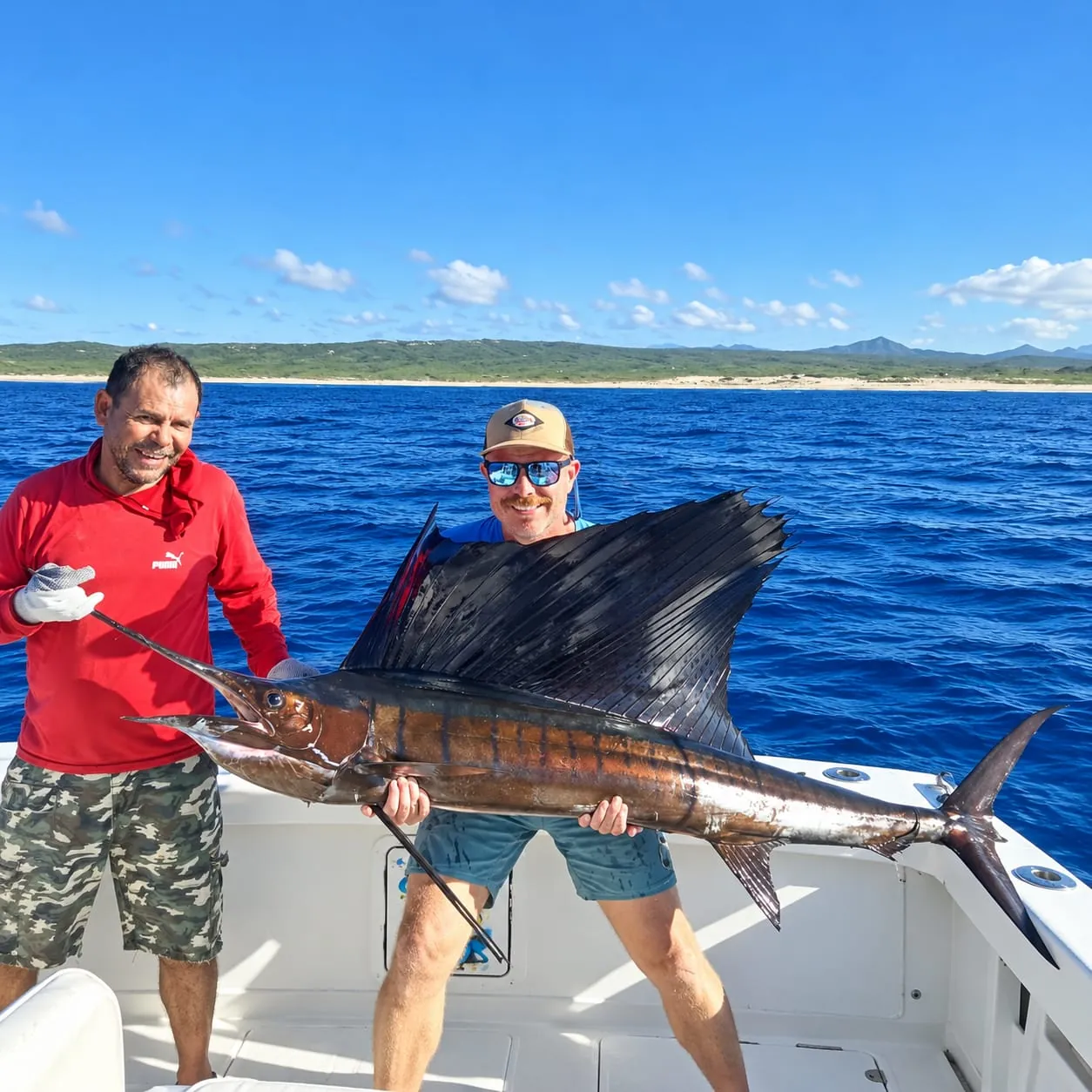 Two anglers with a sailfish, with the coast of Los Cabos behind the boat, October 2026